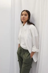 Woman in white shirt and olive green trousers leaning against a white fabric backdrop