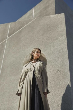 Low-angle shot of a woman in a long tan trench coat and large round hat standing against a tall brutalist concrete architectural wall.