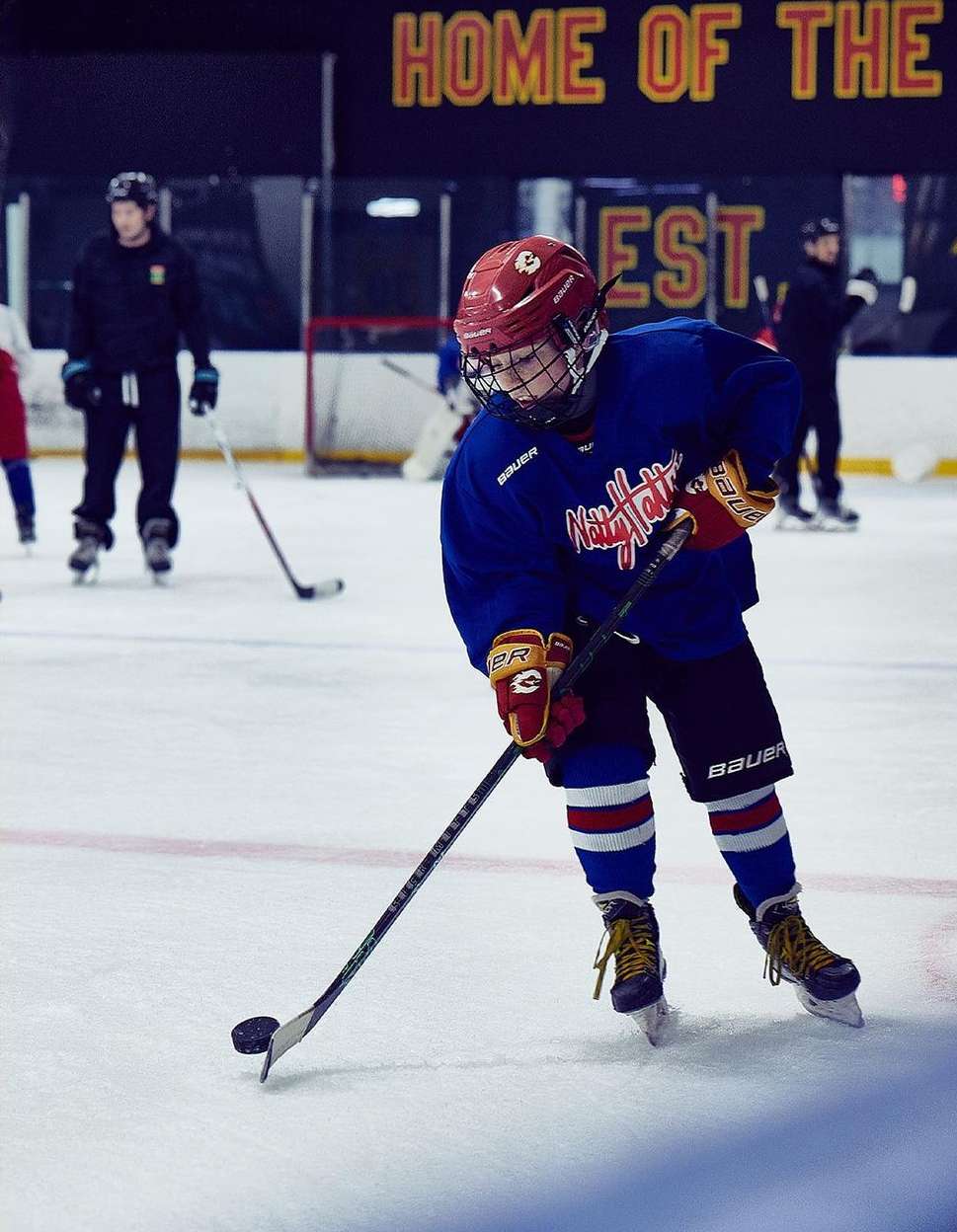 Young hockey player with a stick on the ice rink wearing a helmet, pads, and a team jersey, practicing for a game with coaches.