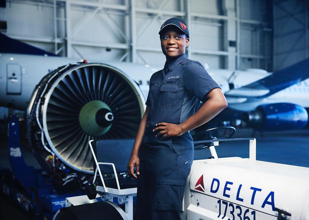 Female aircraft mechanic in uniform stands in hangar, smiling confidently with jet engine and plane in background. Aviation industry.