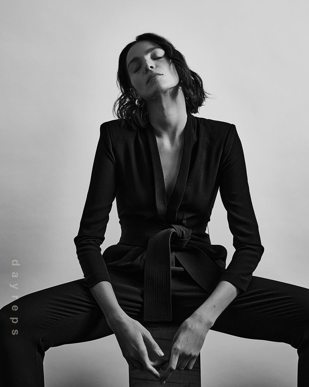 Monochrome portrait of a young adult female with short dark hair wearing a black suit, sitting indoors against a plain background.