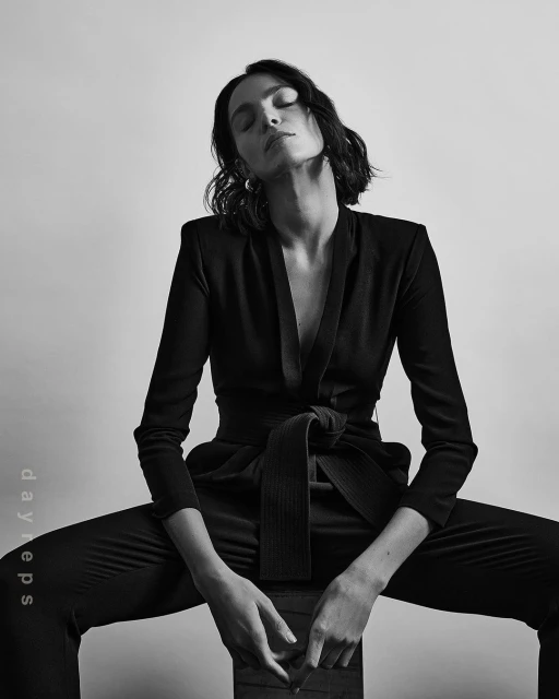 Monochrome portrait of a young adult female with short dark hair wearing a black suit, sitting indoors against a plain background.
