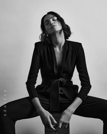Monochrome portrait of a young adult female with short dark hair wearing a black suit, sitting indoors against a plain background.