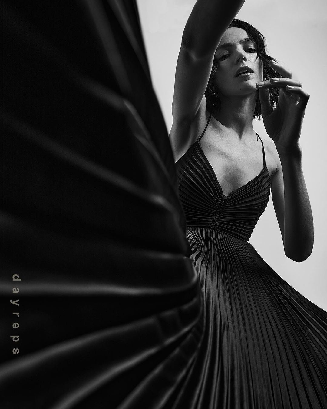 Model in a black pleated dress posing dynamically with dramatic lighting in a monochrome fashion photograph.
