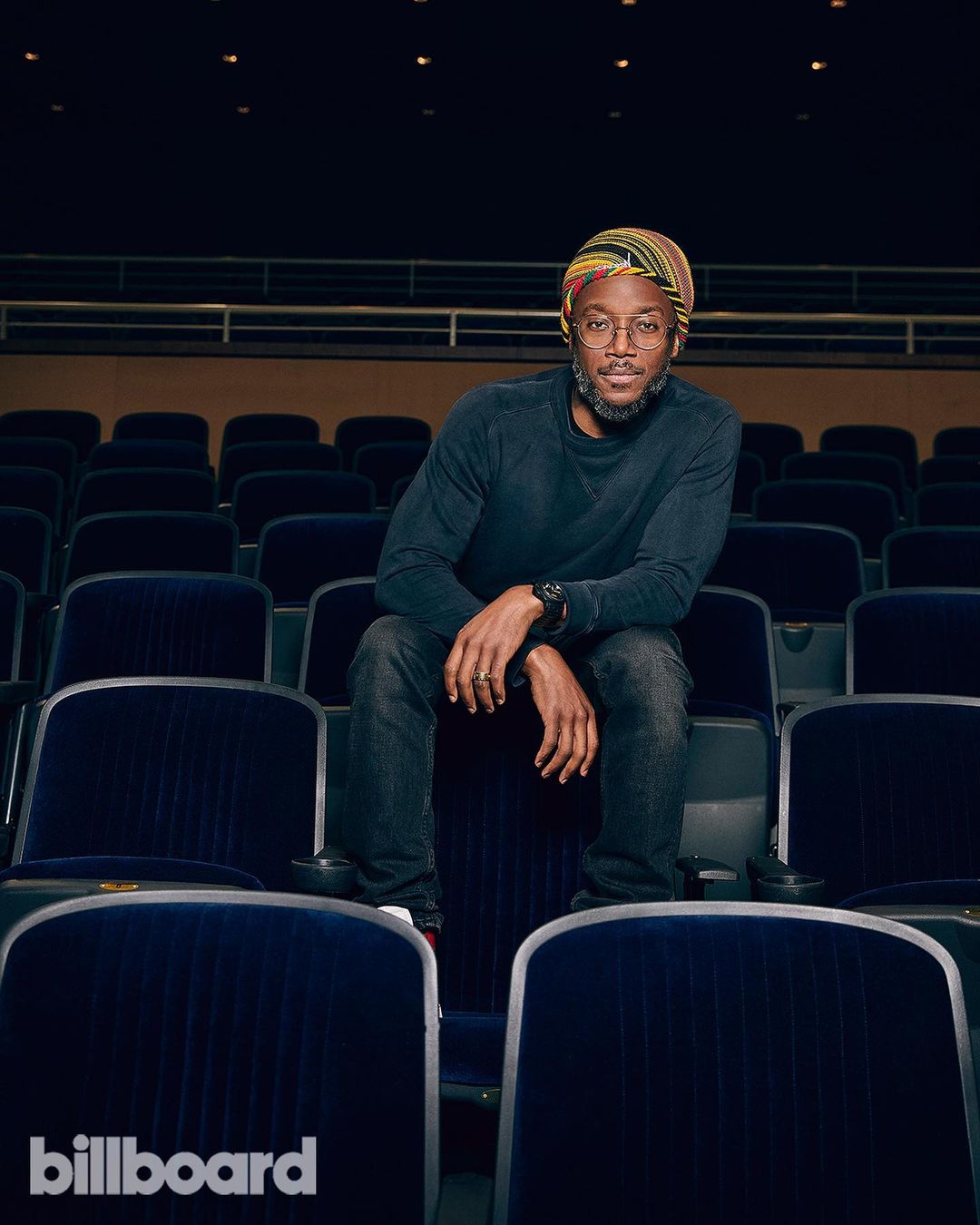 Portrait of a Black adult male sitting in an empty theater with navy blue seating and dark lighting wearing a patterned hat and dark clothing.
