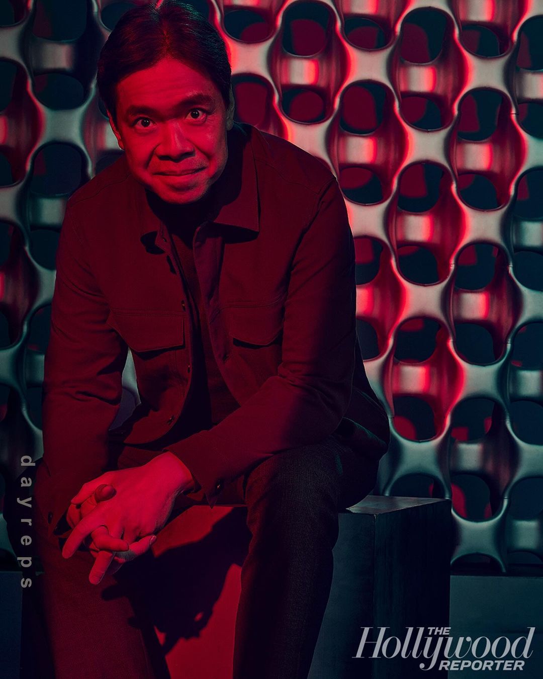 Adult male poses indoors against patterned wall in low light. He sits in dark clothing with his hands clasped together, and has short dark hair.