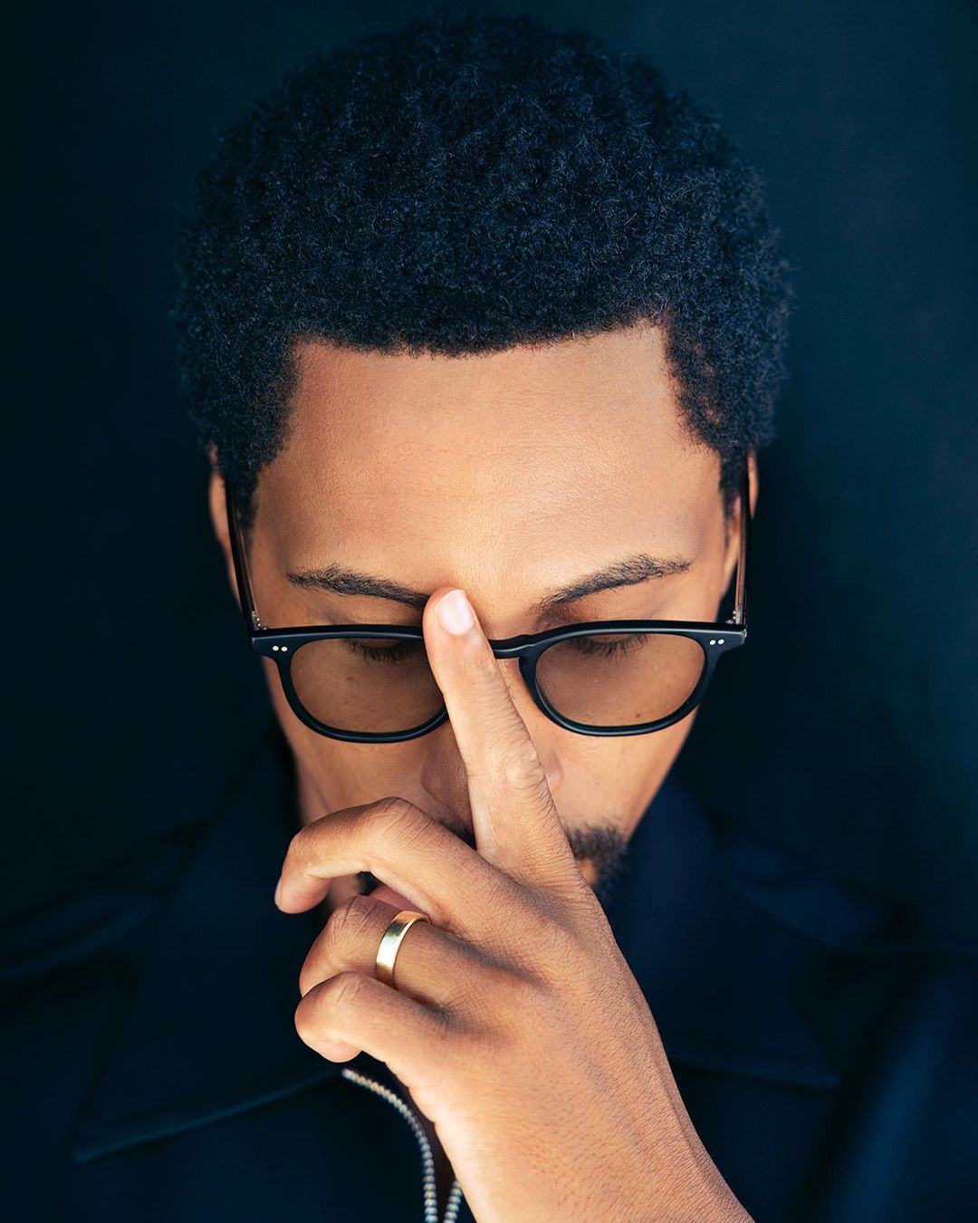 Close-up portrait of a Black adult male wearing glasses, with his finger touching his forehead, against a dark background.