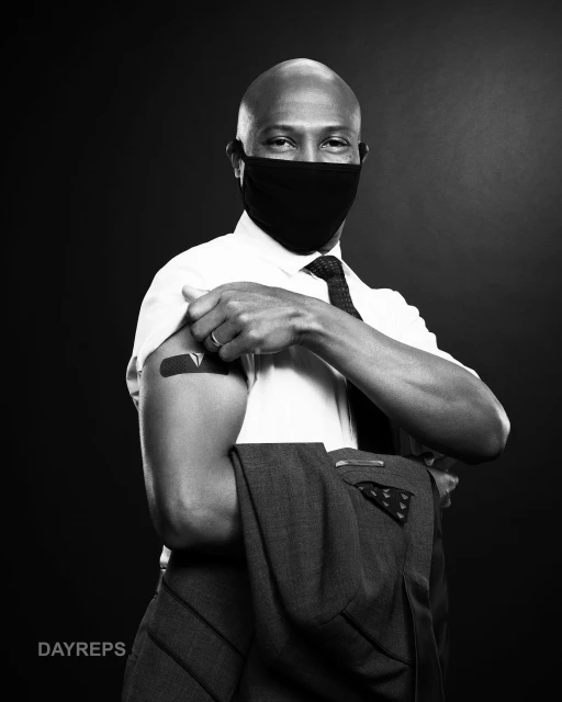 Bald man wearing a black mask, white shirt, and tie, with an adhesive bandage on his arm. He holds a grey jacket in the studio.