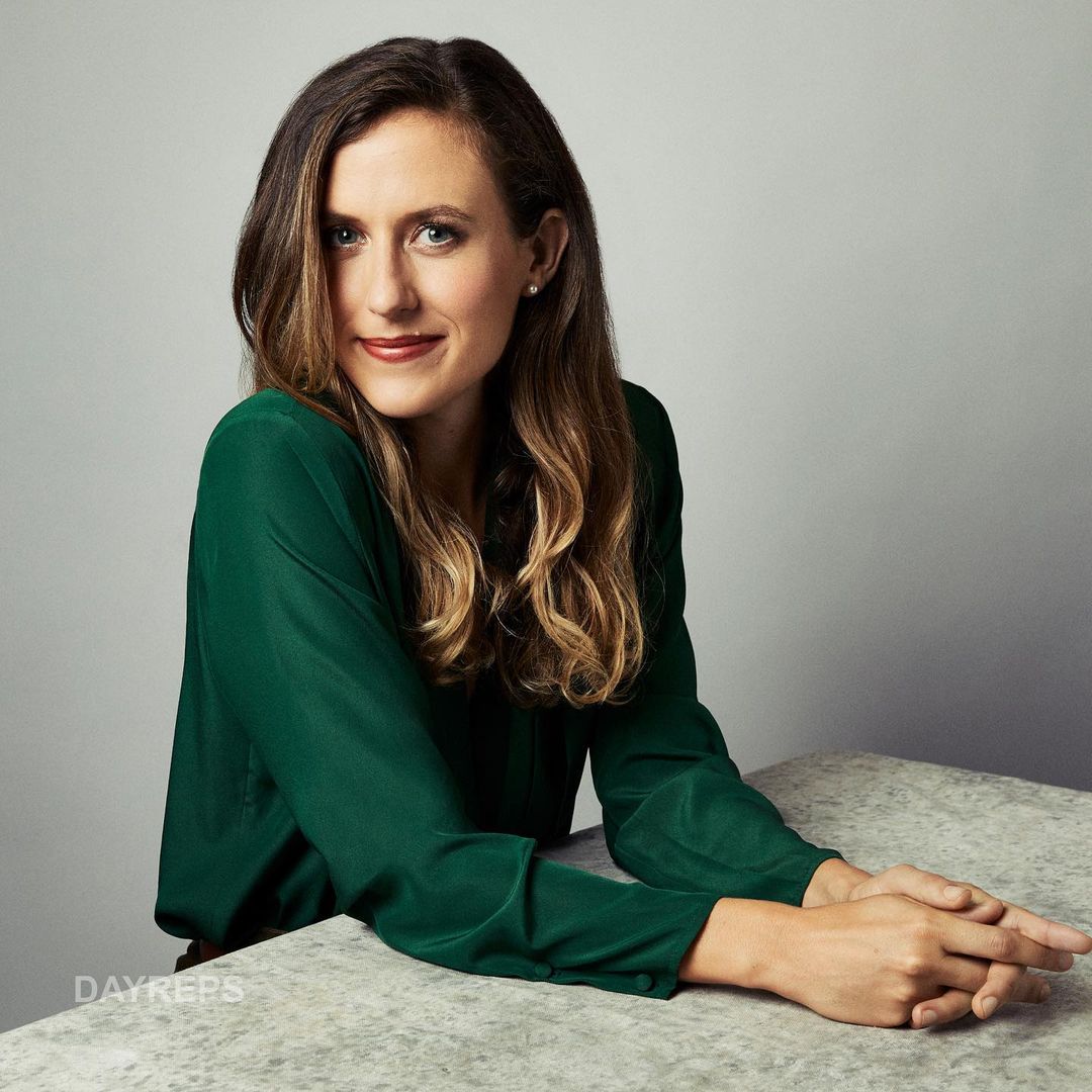 Portrait of a smiling adult woman with long brown hair wearing a green blouse at a table against a grey background in a photo studio.
