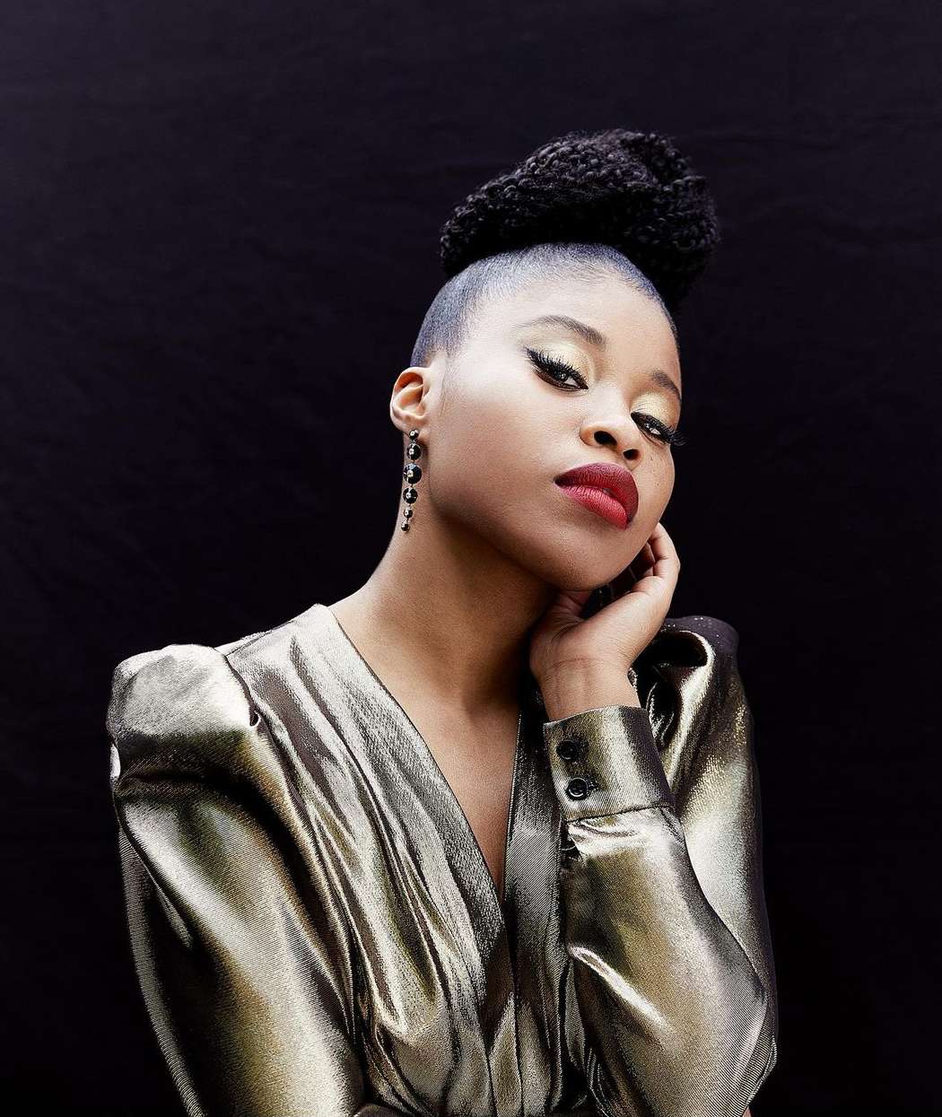 Portrait of an adult woman with dark skin and hair in a high bun, wearing a gold dress with a black background.