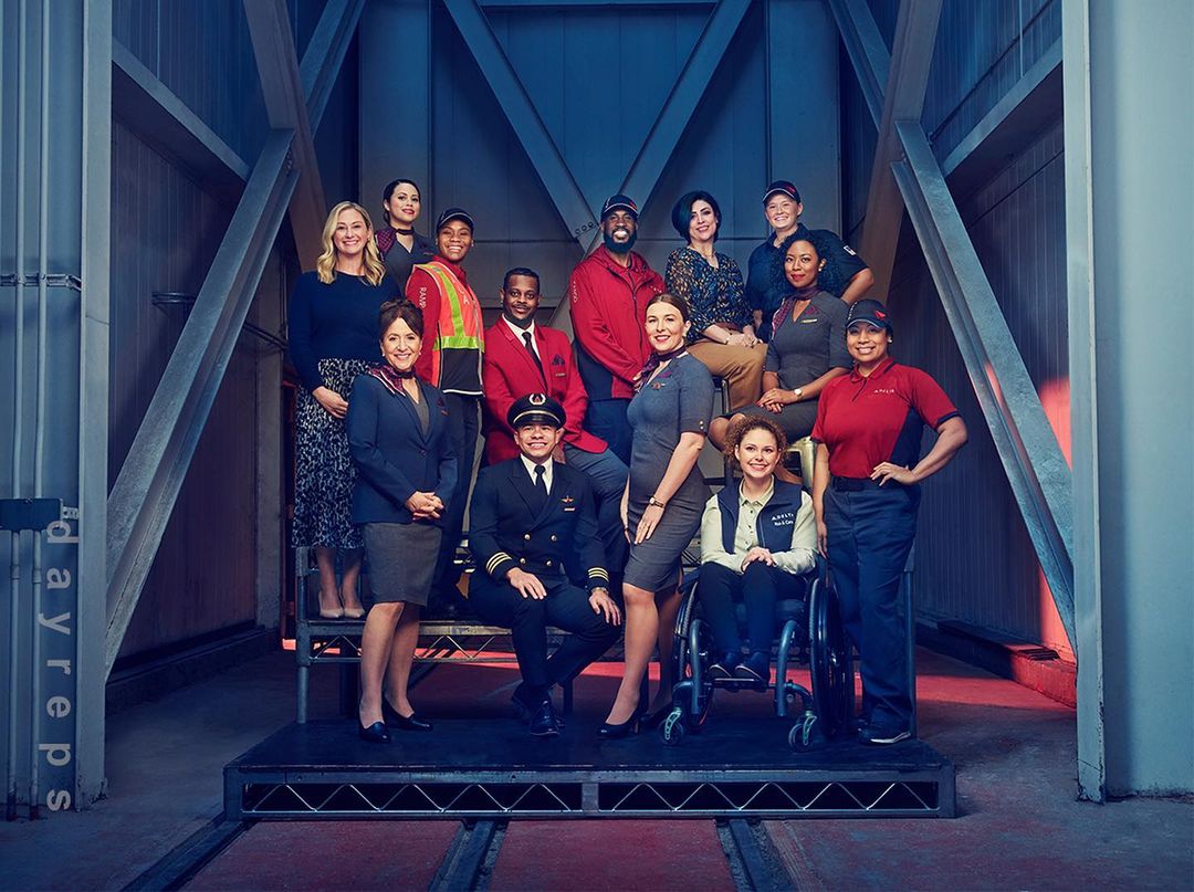 Group portrait of diverse airline employees in uniform posing on a metal platform with an industrial blue and grey background, looking friendly.