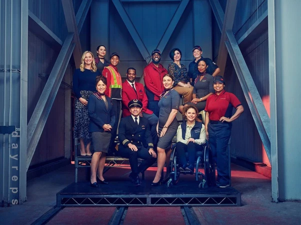 Group portrait of diverse airline employees in uniform posing on a metal platform with an industrial blue and grey background, looking friendly.