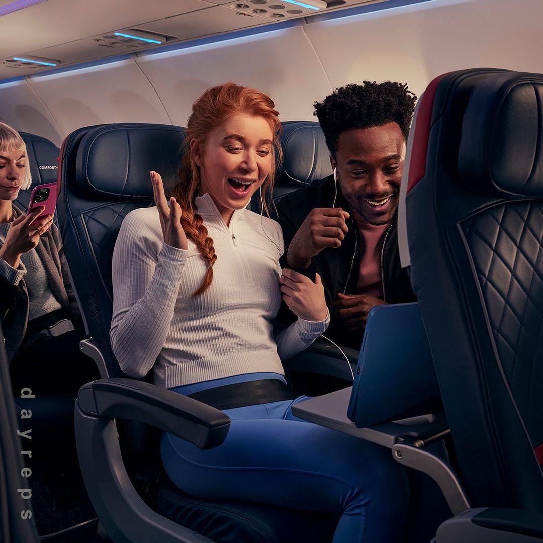 Three people on an airplane are watching a laptop. Two people are looking at the screen with smiling expressions.