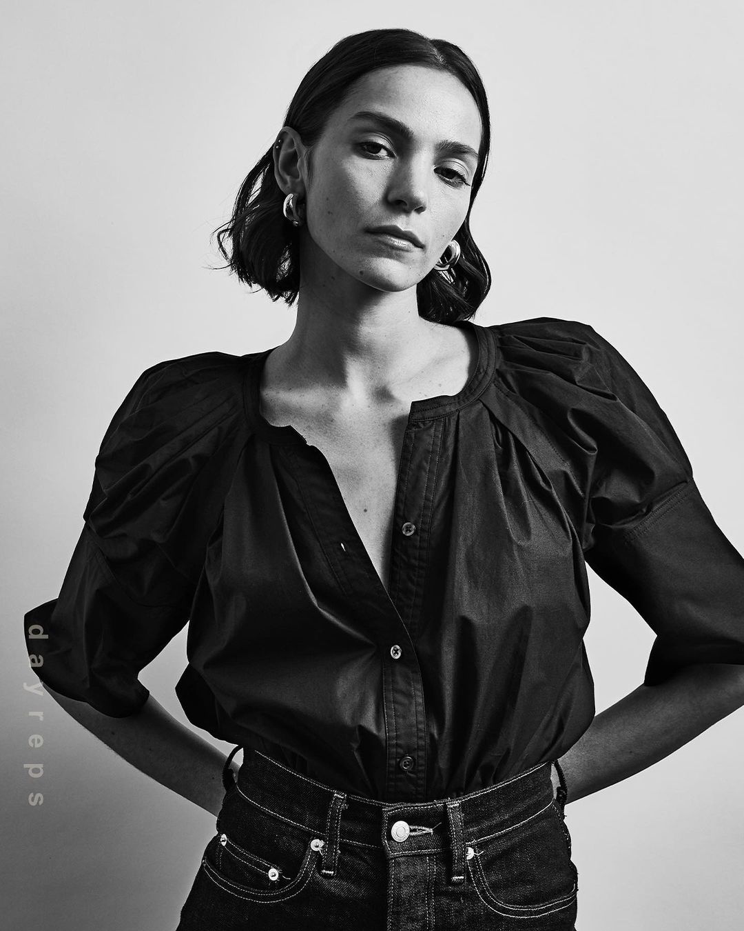 Monochrome portrait of a woman in a dark blouse and jeans, standing against a neutral backdrop with hands behind her.