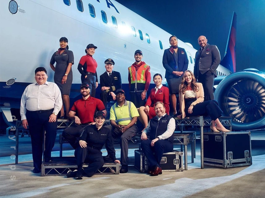 Group of Delta Airlines employees in uniform posing in front of a passenger jet, including pilots, flight attendants, and ground crew.