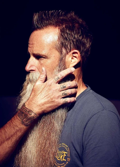 Portrait of a bearded man with a long beard being touched on the face, wearing a t-shirt, tattoos are visible on his arm and fingers.