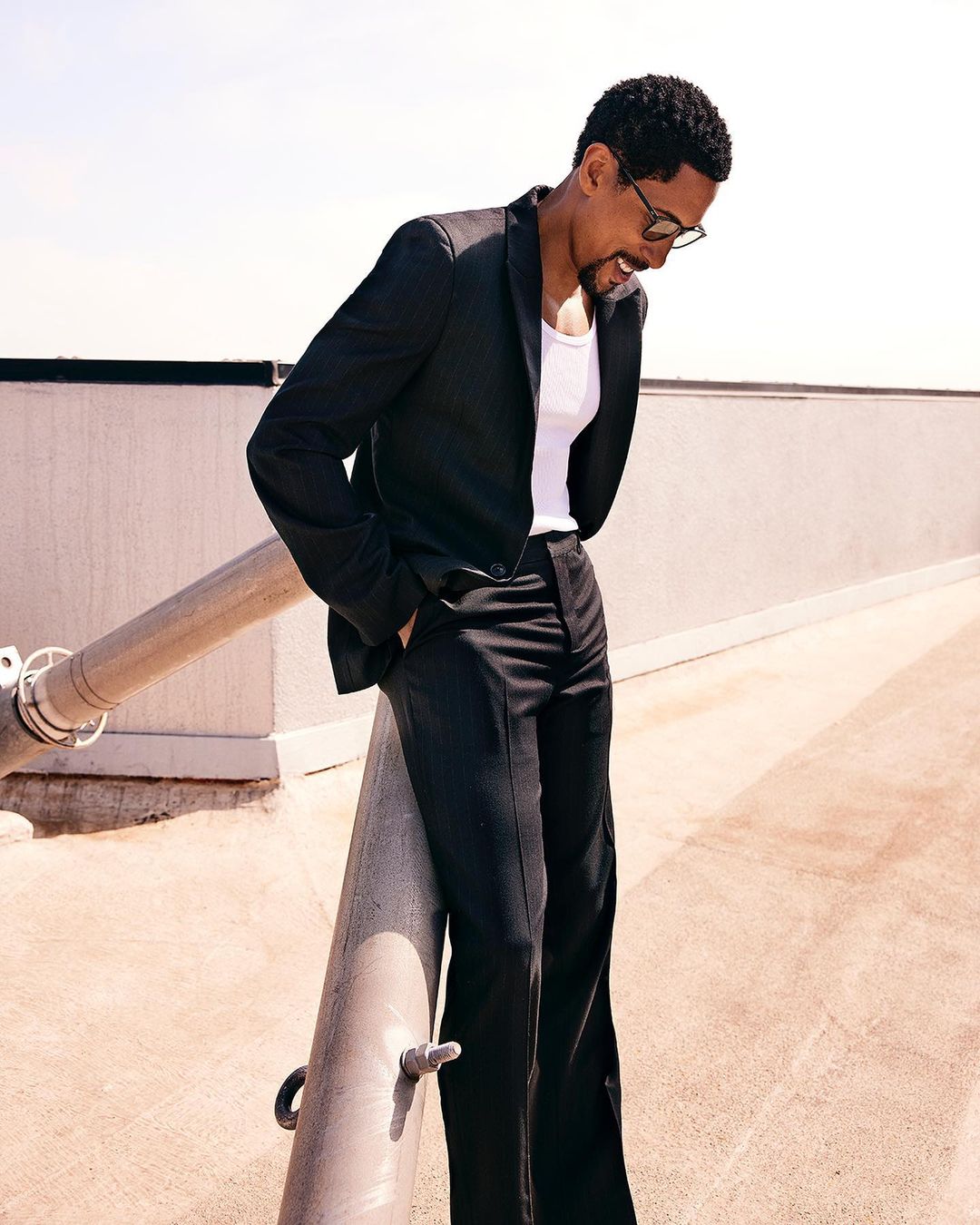 Man in a black suit and glasses leans against a pipe outdoors under bright sunlight, with a neutral wall visible in the background.
