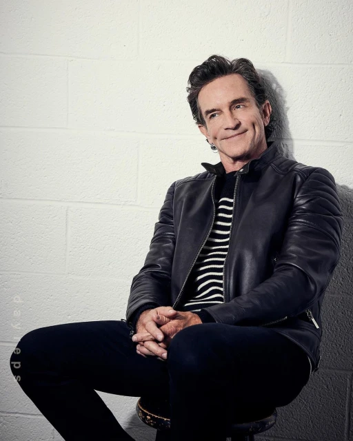 Portrait of middle-aged adult male with dark hair wearing a black leather jacket and striped shirt, sitting against a white brick wall.