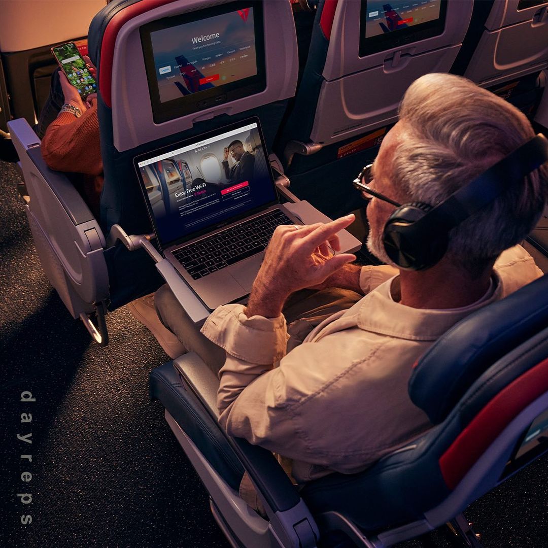 Mature adult male sits inside an airplane cabin with a laptop and headphones, browsing the internet mid-flight, with another passenger.