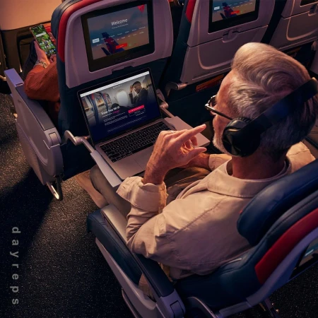 Mature adult male sits inside an airplane cabin with a laptop and headphones, browsing the internet mid-flight, with another passenger.