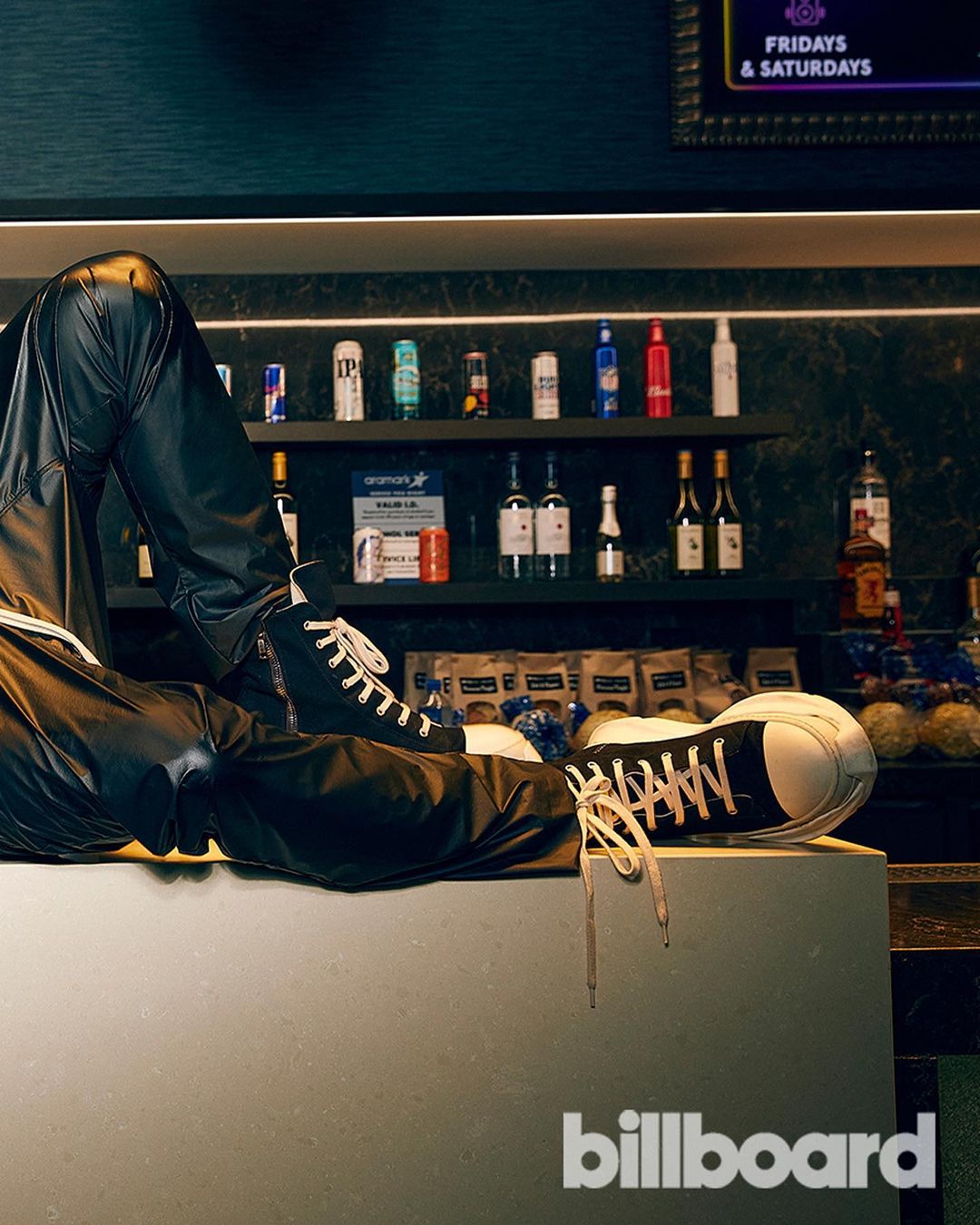 A pair of sneakers worn with leather pants are posed on a counter with an out-of-focus liquor shelf and the Billboard logo in the foreground.
