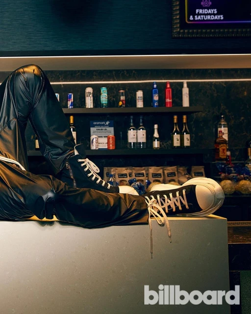 A pair of sneakers worn with leather pants are posed on a counter with an out-of-focus liquor shelf and the Billboard logo in the foreground.