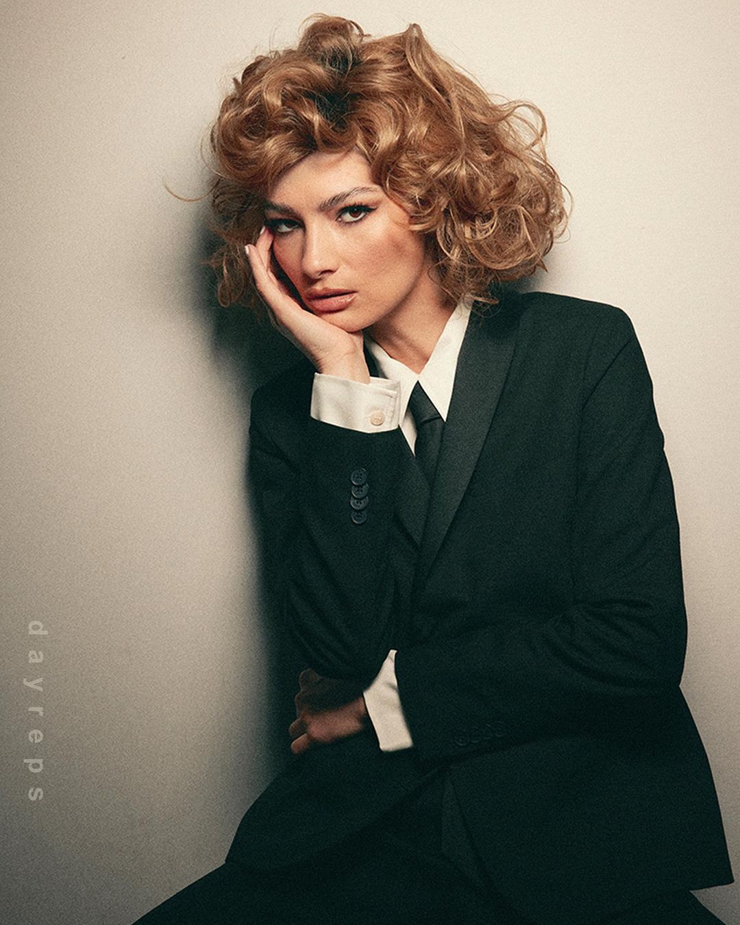 Elegant woman in a black suit and tie with curly hair poses against a muted wall, looking pensively at the camera with hand on face.