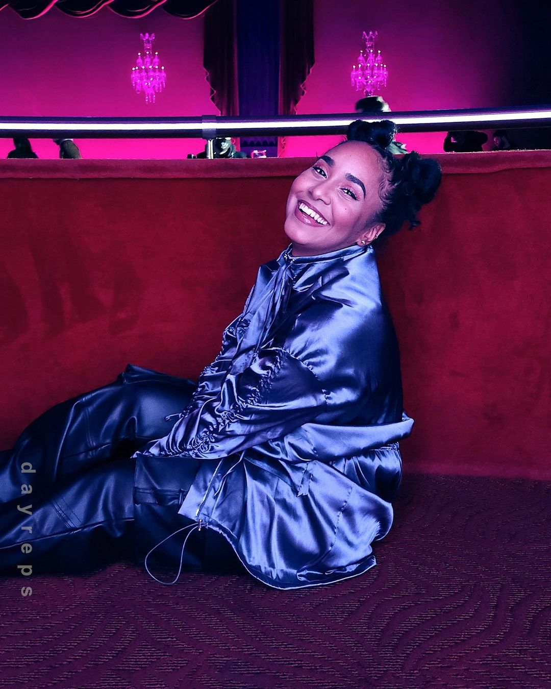 Smiling woman with updo in satin jacket and pants sits in a theatre box with red velvet couch under chandeliers and magenta lighting.