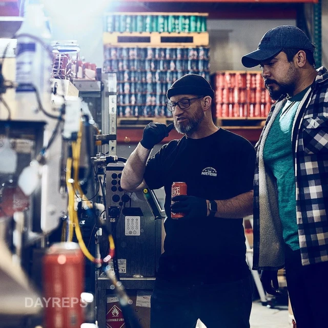 Two men at a beverage canning facility. One man in a beanie holds a canned drink. Pallet of cans and machinery are visible in background.
