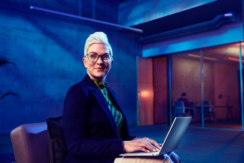 Professional woman with short white hair, glasses, and a dark blazer sits in a modern office while using a laptop.
