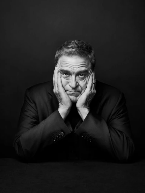 Monochrome portrait of a mature adult male with hands on face wearing a suit jacket against a dark background in eye-level view.