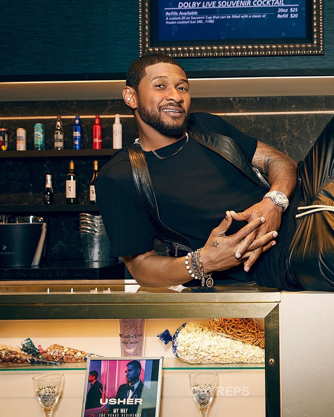 Usher poses in a black vest and pants, leaning on a counter with drinks and snacks, showcasing a casual yet stylish look indoors.