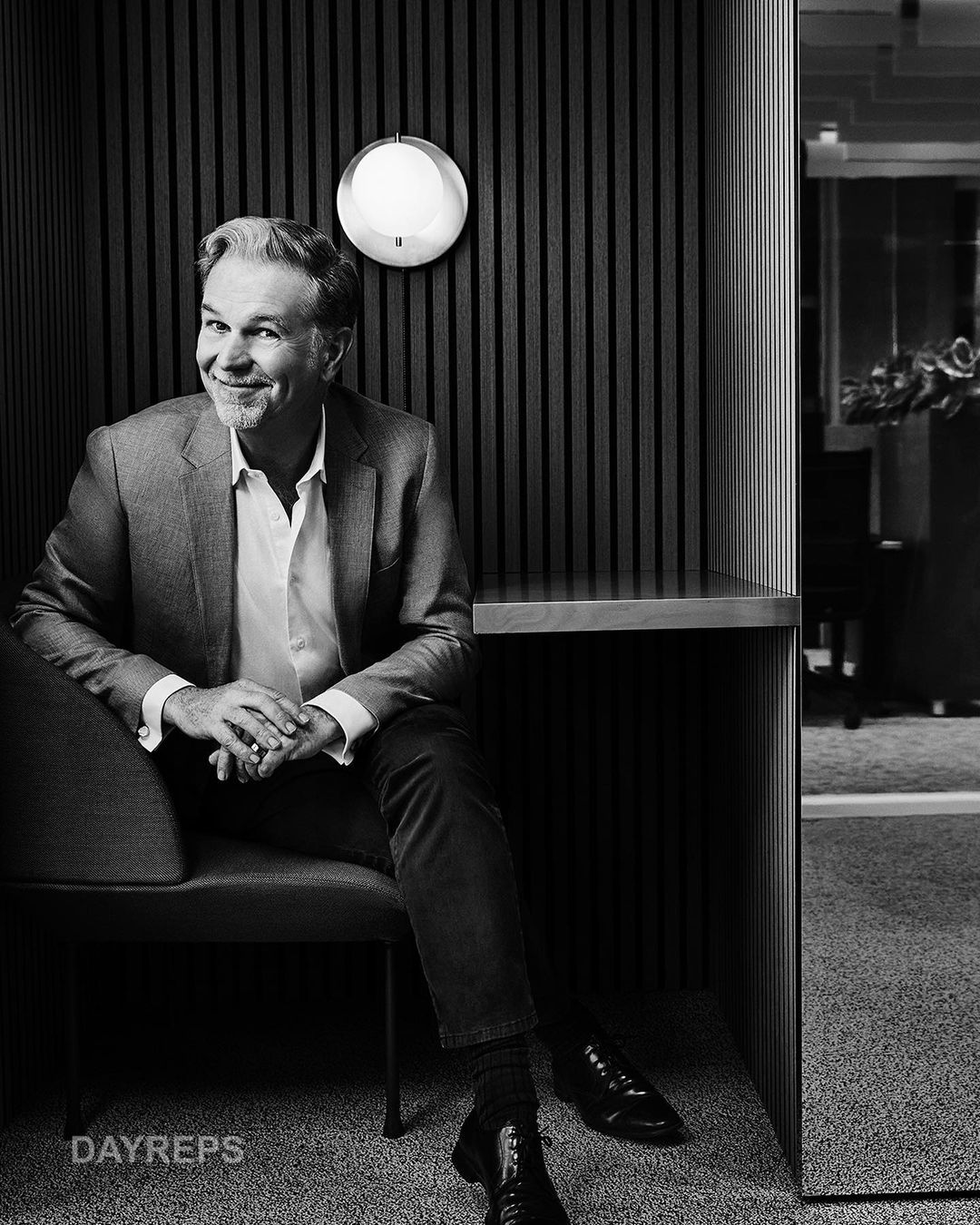 Portrait of a grey-haired man in a grey suit sitting indoors in a black chair against a wood-paneled wall with a spherical light.