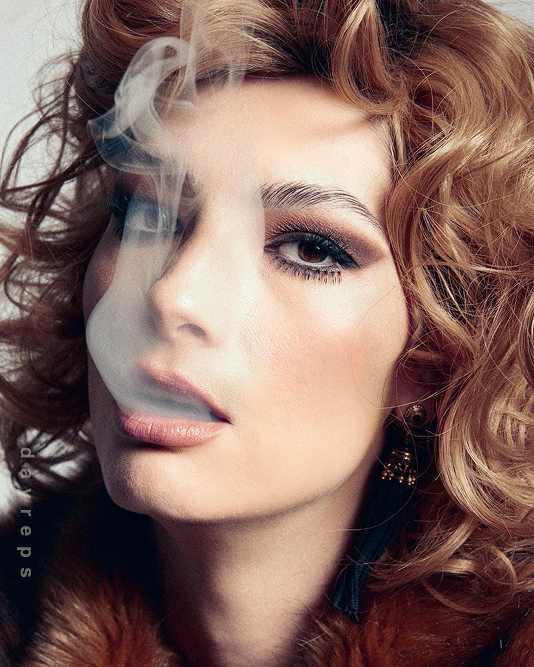 Glamorous adult woman in fur coat with curled hair posing with smoke against a light background, styled with statement earrings.