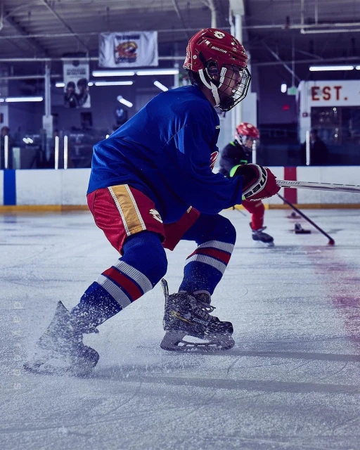 Action shot of a young hockey player in full gear skating on an ice rink, wearing a red helmet, blue jersey and red shorts.