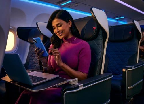 A woman sits in a comfortable airplane seat, using her smartphone and laptop, showcasing the blend of travel and technology.