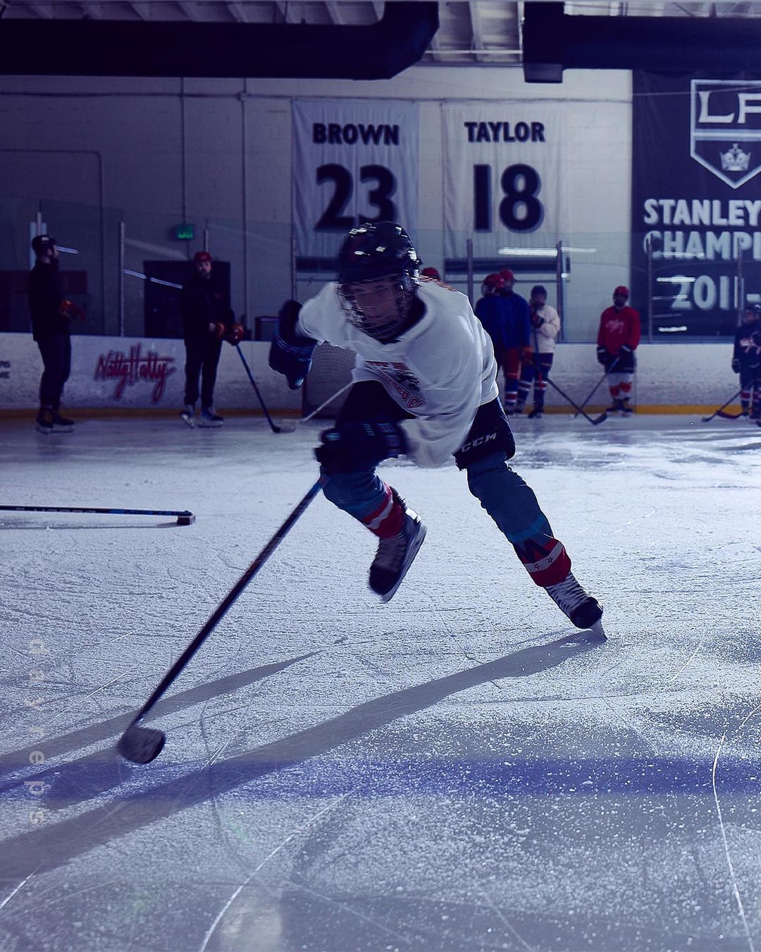 A hockey player skates across an ice rink, stick in hand, illuminated by overhead lights in a full shot indoors.