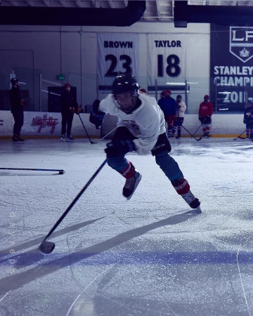 A hockey player skates across an ice rink, stick in hand, illuminated by overhead lights in a full shot indoors.