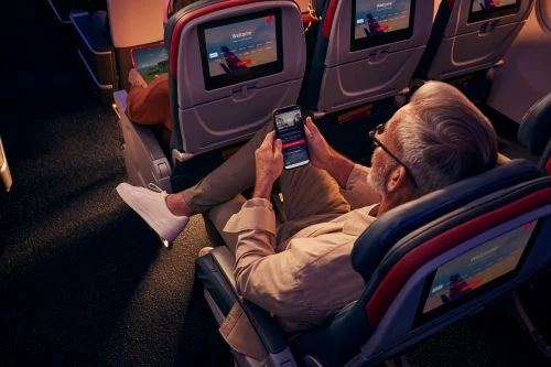 Senior man relaxing on an airplane, wearing glasses, using his smartphone, and wearing white sneakers, surrounded by seating.