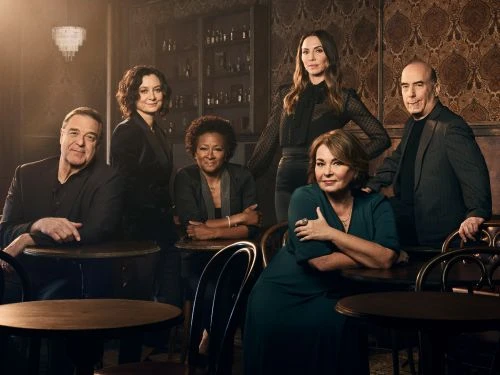 Group portrait of six actors indoors at a bar or restaurant, well-lit with visible bottles and glasses behind them.