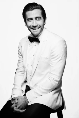 Black and white portrait of a smiling adult male in a white tuxedo with a black bow tie, seated on a stool against a white background.
