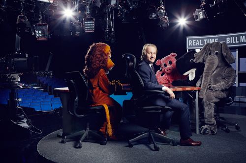 Bill Maher sits at a desk in a television studio with a lion, pig, steer, and elephant in costume, with studio lighting in the background.
