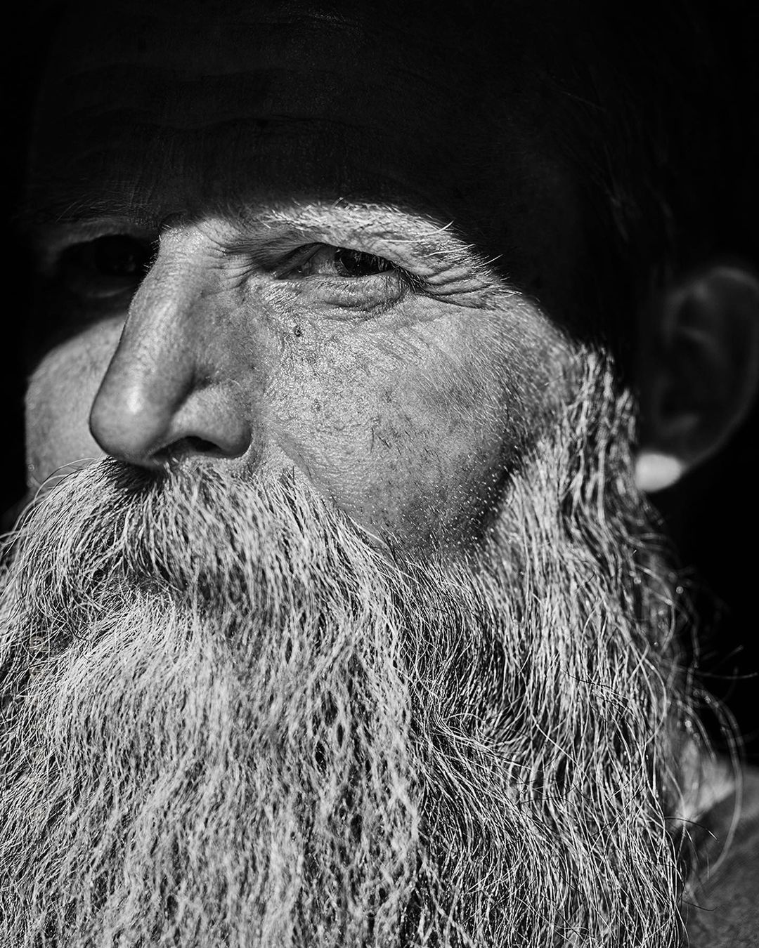 Close-up black and white portrait of an elderly man with a long textured beard and serious expression by Christopher Patey