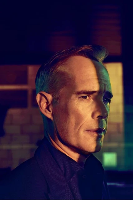 Portrait of an adult man with short hair. His face is partially lit with blue and green hues against a dark textured background.