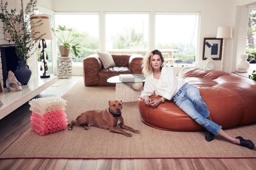 Woman in a white shirt and blue jeans lounging on a brown beanbag chair in a bright living room with a dog lying on the rug.