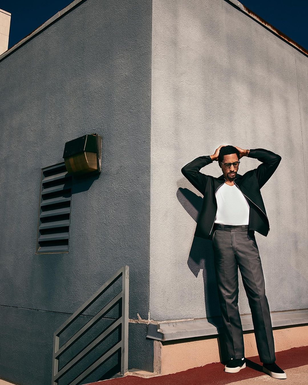Stylish adult male in jacket and trousers poses outdoors with hands on head against a grey building in urban setting with cool lighting.