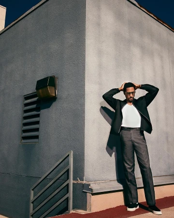 Stylish adult male in jacket and trousers poses outdoors with hands on head against a grey building in urban setting with cool lighting.