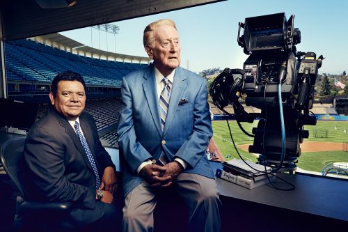 Two men in suits sitting near a large television camera at a baseball stadium, with blue stadium seats visible in the background.