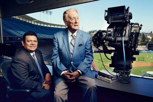 Two men in suits sitting near a large television camera at a baseball stadium, with blue stadium seats visible in the background.