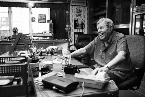 Black and white photo of an older man sitting at a cluttered desk in a wood-paneled office, smiling, with natural light from a window.